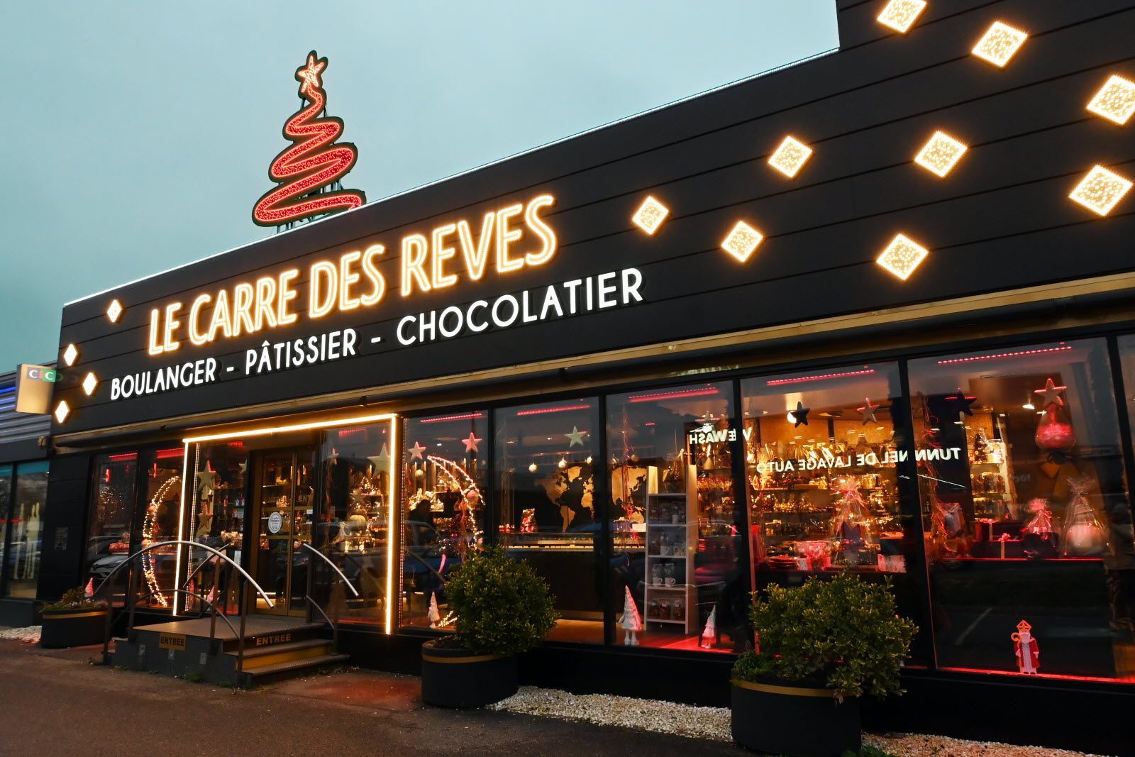 Extérieur de la boulangerie-chocolaterie Le Carré des Rêves la nuit, décoré de sapins de Noël illuminés et de guirlandes lumineuses.