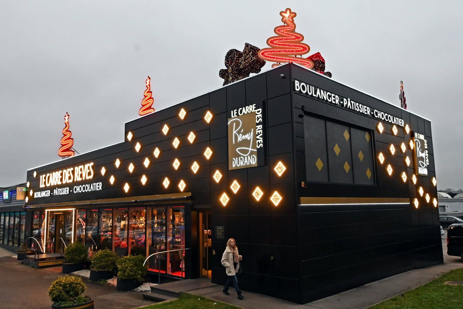 Boulangerie noire avec éclairage d'accentuation doré et décorations de sapin de Noël sur le toit, sous un ciel nuageux.