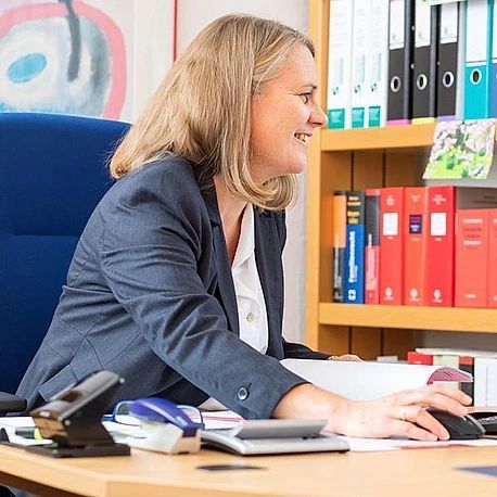 Eine Frau sitzt an einem Schreibtisch vor einem Bücherregal und benutzt eine Computermaus.