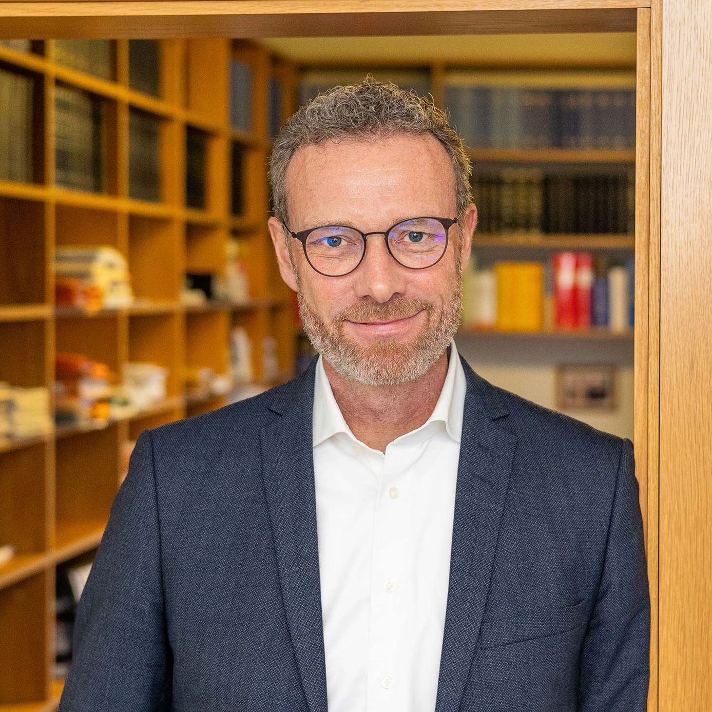 Ein Mann in Anzug und Brille steht vor einem Bücherregal.