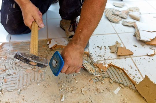 Un hombre está arrodillado en el suelo sosteniendo un martillo y una espátula.