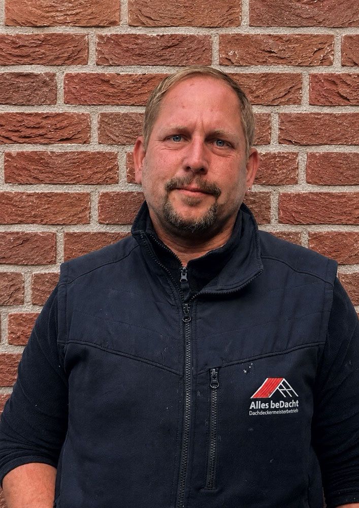 Ein Mann mit Hut und Jacke steht vor einer Backsteinmauer.