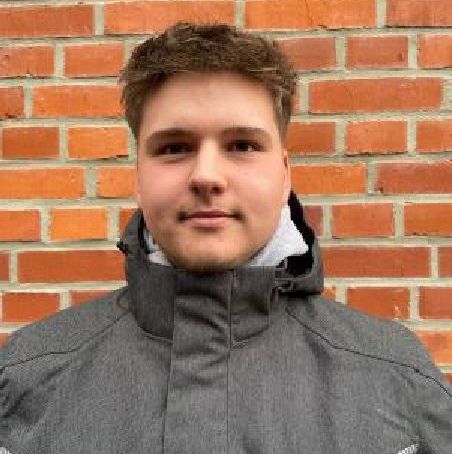 Ein junger Mann in einer grauen Jacke steht vor einer Backsteinmauer.