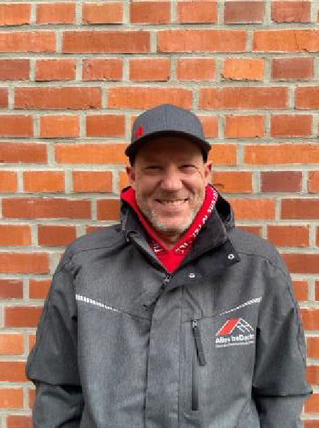 Ein Mann mit Hut und Jacke steht vor einer Backsteinmauer.