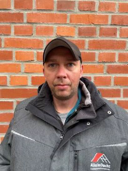 Ein Mann in Jacke und Hut steht vor einer Backsteinmauer.