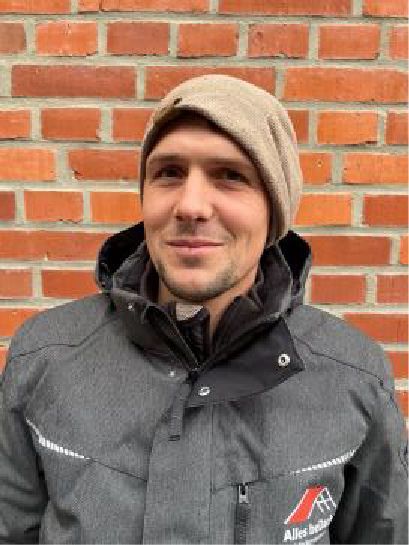 Ein Mann mit Hut und Jacke steht vor einer Backsteinmauer.