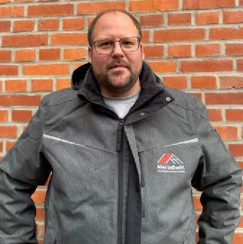 Ein Mann mit Brille und Jacke steht vor einer Backsteinmauer.