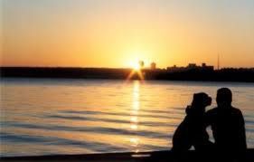 Silhouette of a person and dog watching sunset over water, skyline in the distance.