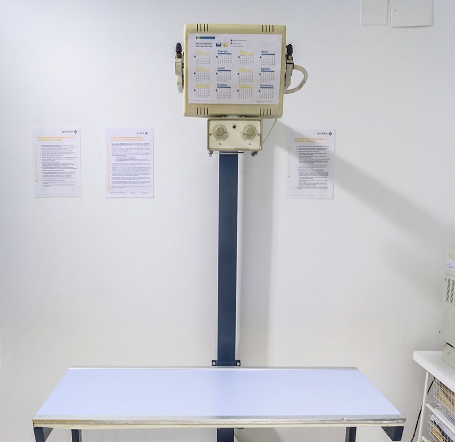 Máquina de rayos X sobre una mesa de examen blanca en una clínica, con carteles de instrucciones en la pared.