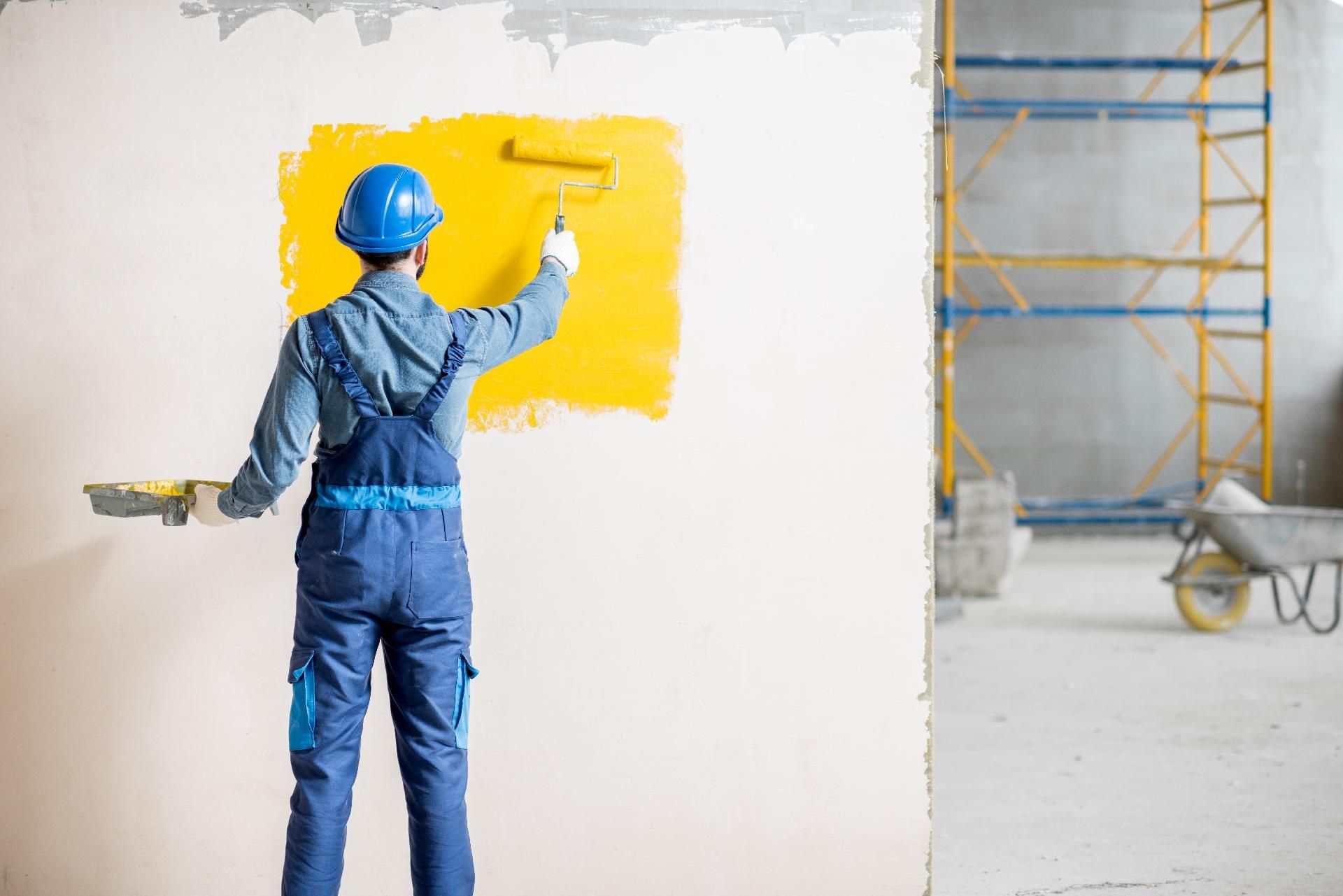 Ein Arbeiter in blauem Overall und Schutzhelm streicht auf einer Baustelle eine Wand gelb.