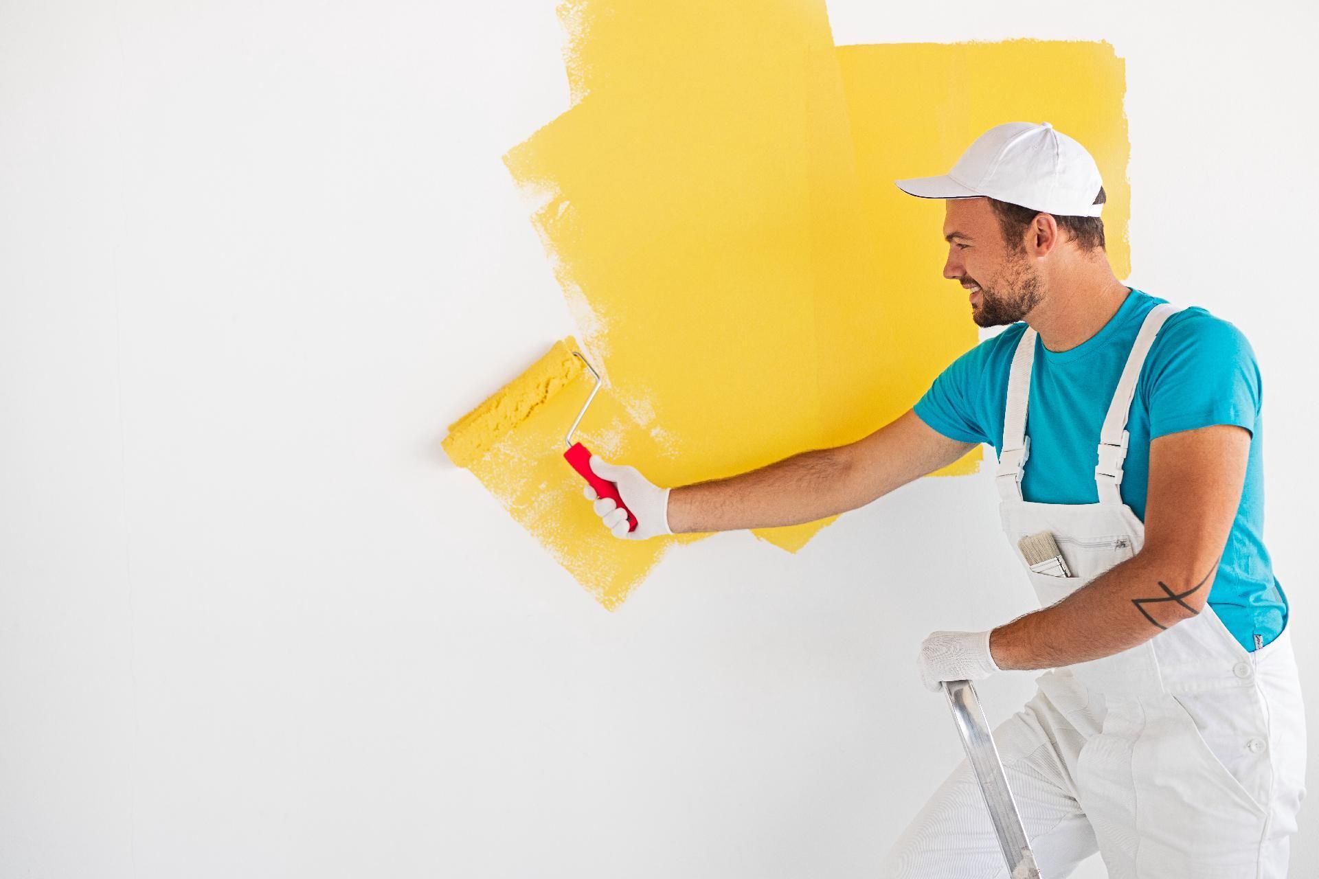 Ein Maler rollt leuchtend gelbe Farbe auf eine weiße Wand neben einer Leiter.