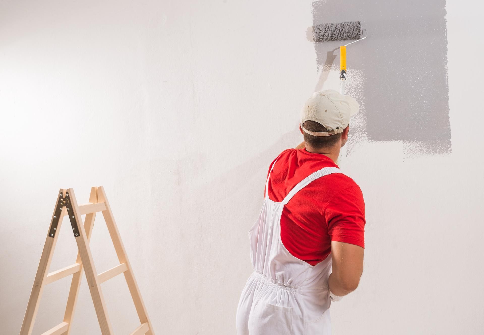 Eine Person streicht neben einer Leiter mit einer Farbrolle eine Wand weiß.