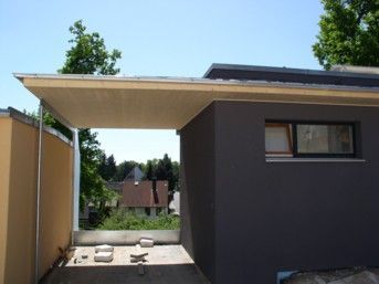 Ein Carport mit hellem Dach ist an ein dunkelgraues Gebäude mit Fenster angebaut
