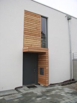 Moderner Hauseingang mit Holzelementen, grauer Tür, hohem Fenster und Briefkasten. 