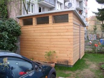 Ein hölzerner Geräteschuppen mit Fenstern steht neben einem Auto.