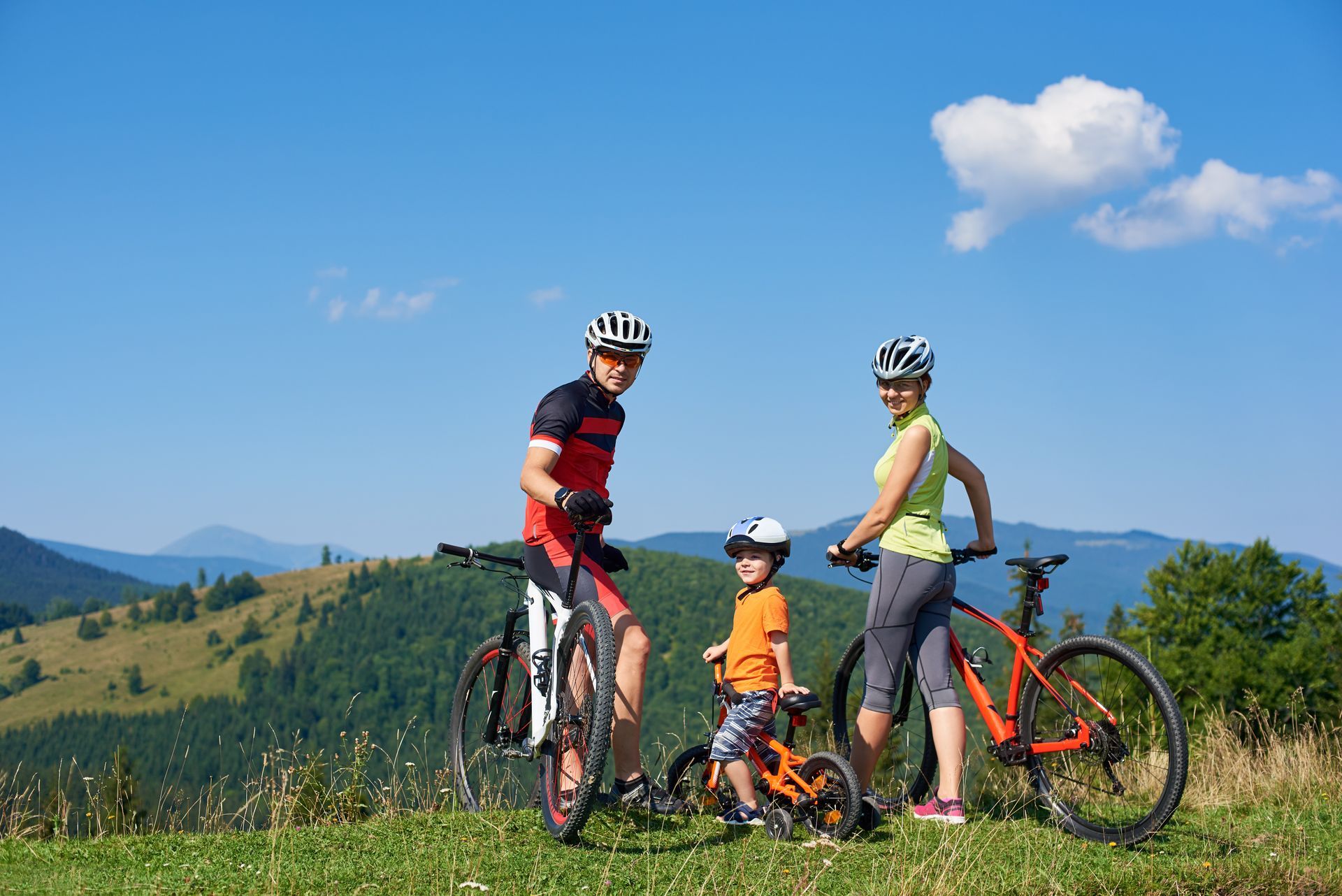 Drei Radfahrer mit Fahrrädern auf einem grasbewachsenen Hügel unter blauem Himmel und Bergen.