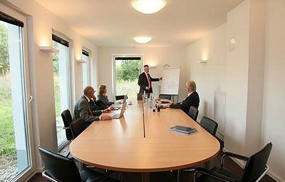 Besprechungsraum mit vier Personen an einem langen Holztisch, eine Person präsentiert an einem Flipchart.