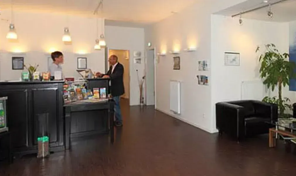 Moderne Lobby mit Rezeption, einem Mann, der mit Angestellten spricht, und schwarzen Sitzgelegenheiten entlang einer weißen Wand