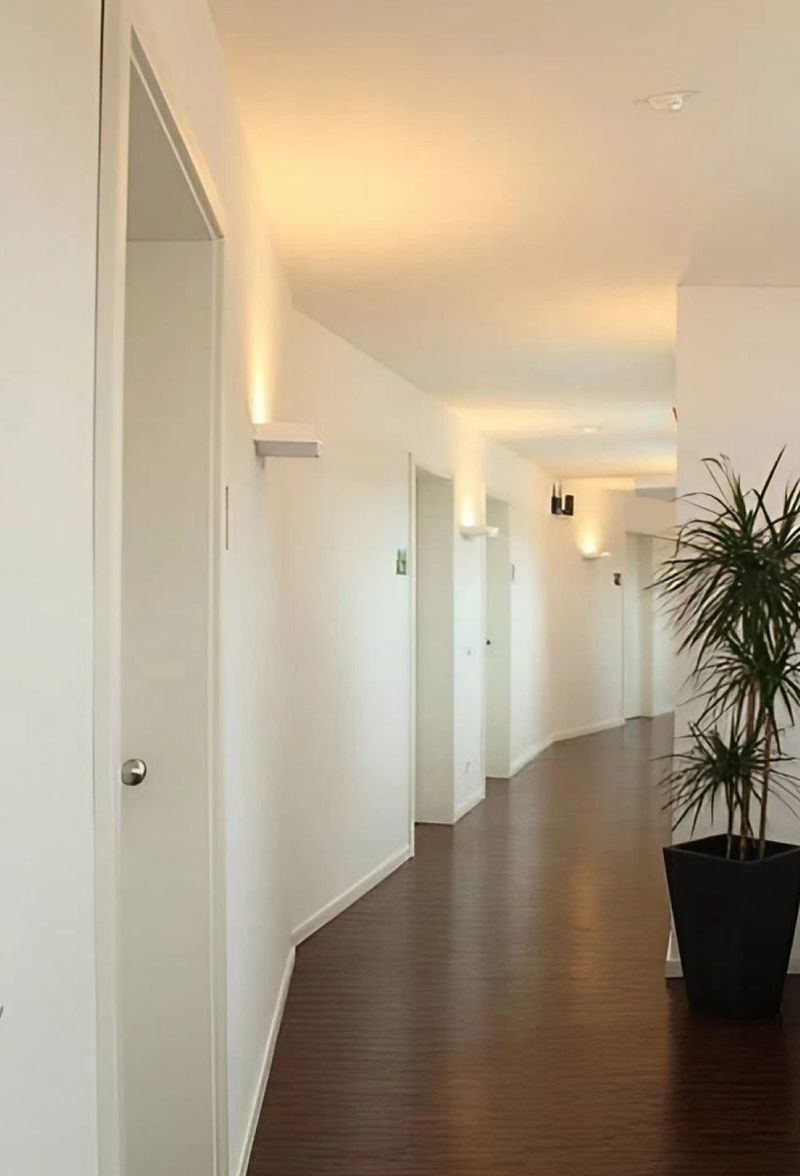 Weißer Flur mit Holzboden, Wandleuchten, mehreren Türen und einer Topfpflanze am Ende.