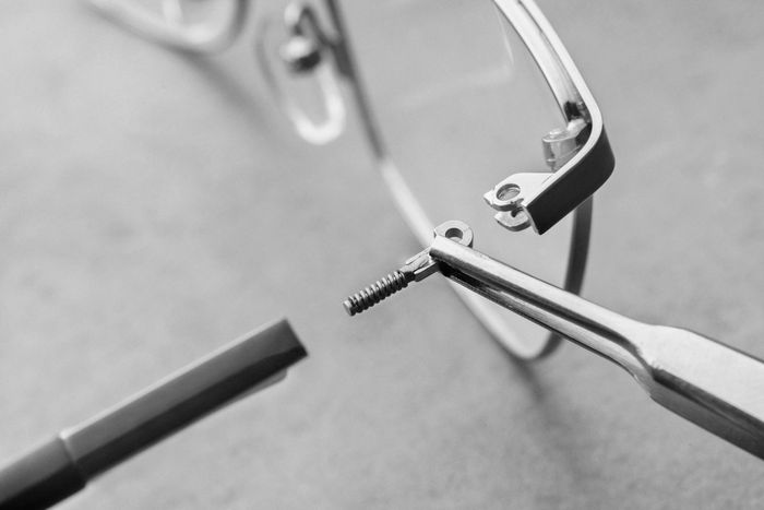 Nahaufnahme einer Brille, die mit einer Zange und einer kleinen Schraube auf einer Arbeitsfläche repariert wird