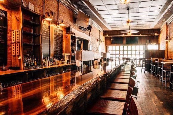 A long bar with copper counter and wooden stools in a dimly lit bar with brick walls.