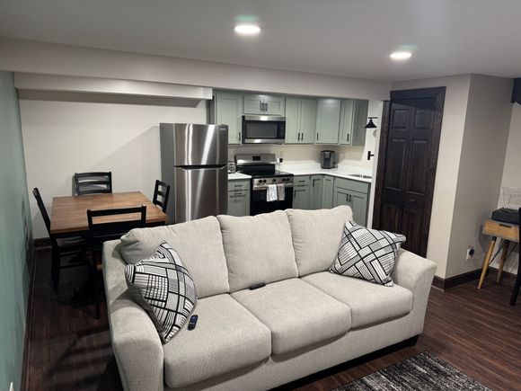 A small apartment with a gray couch, kitchen, and dining table. The kitchen has light green cabinets.