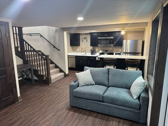 A small living space with a kitchen and stairs. Gray sofa faces the kitchen with a stainless steel fridge.