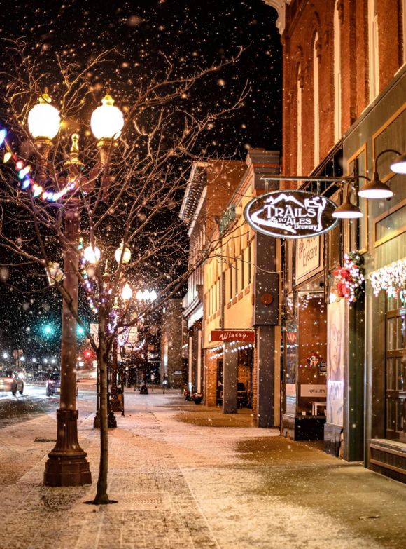 Snowy street with lit storefronts and lampposts at night.