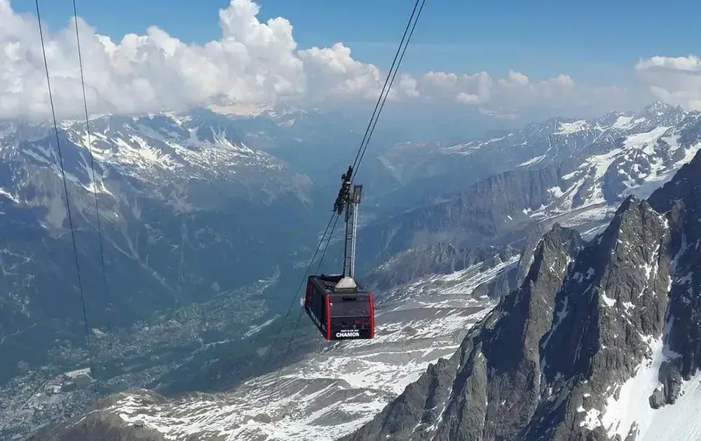 Cable car ascending over a snow-capped mountain range under a blue sky.