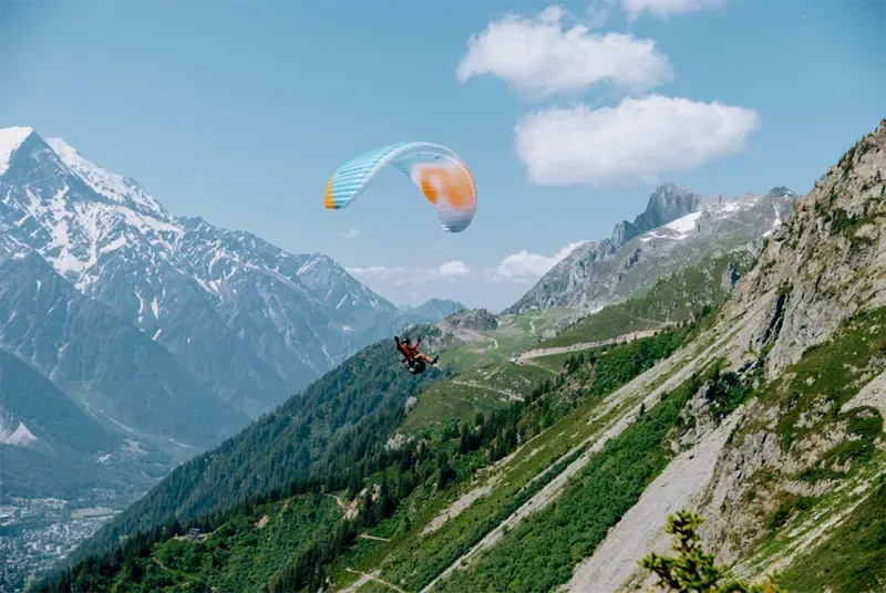 Paraglider soaring over a mountain range with snowy peaks and a lush green hillside on a sunny day.