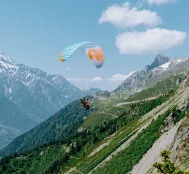 Paraglider soaring over a green mountain range, under a blue sky with fluffy clouds.