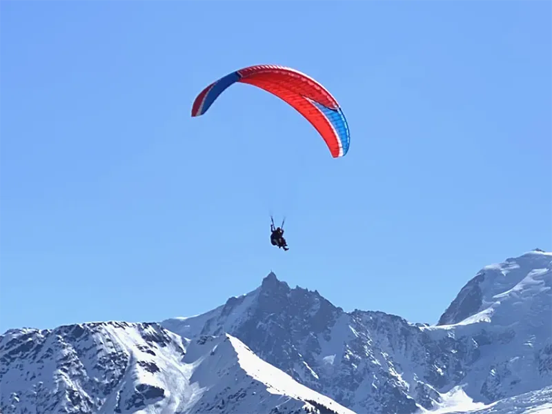 Paraglider soaring above snow-covered mountains under a clear blue sky; colorful red and blue canopy.