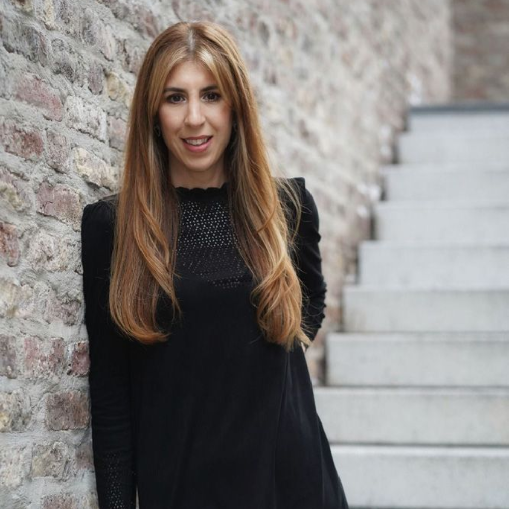 Eine Person mit langen blonden Haaren, die ein schwarzes Kleid trägt, lehnt an einer strukturierten Steinmauer neben einer Betontreppe.