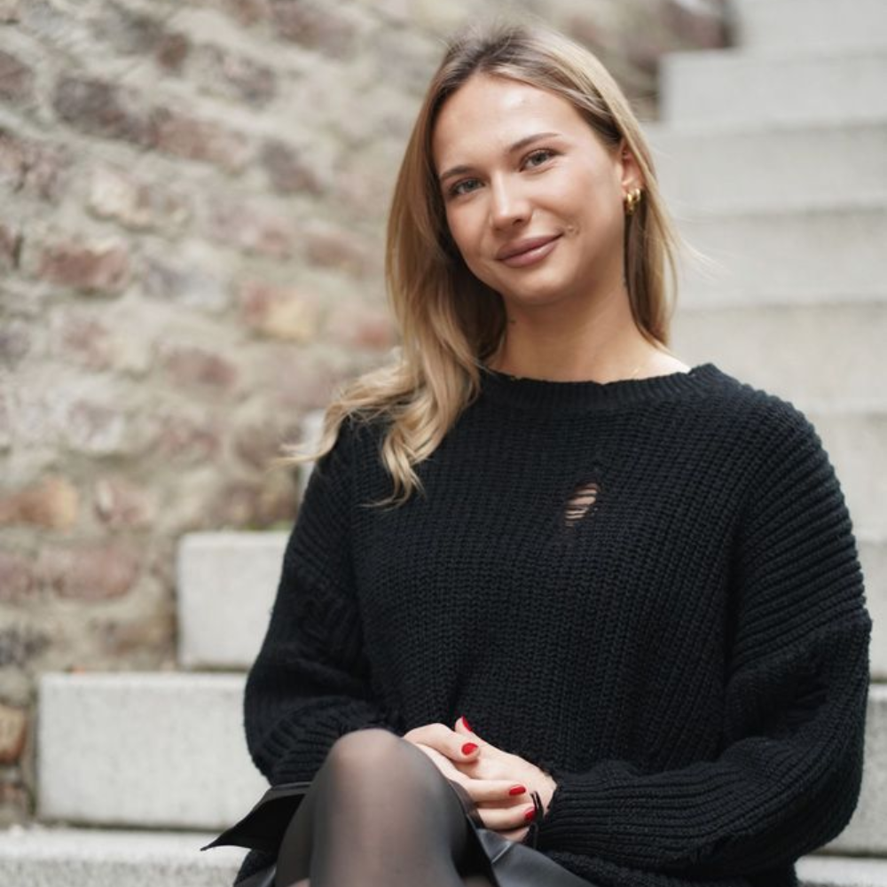 Eine Person mit schulterlangem blonden Haar, die einen schwarzen Pullover trägt, sitzt auf einer Betontreppe vor einer Backsteinmauer.