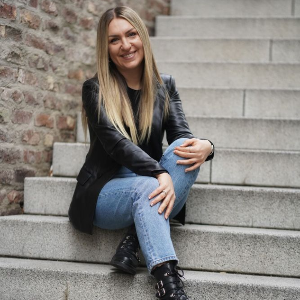 Eine lächelnde Person mit langen blonden Haaren, die einen schwarzen Blazer und Jeans trägt, sitzt auf einer Steintreppe im Freien.