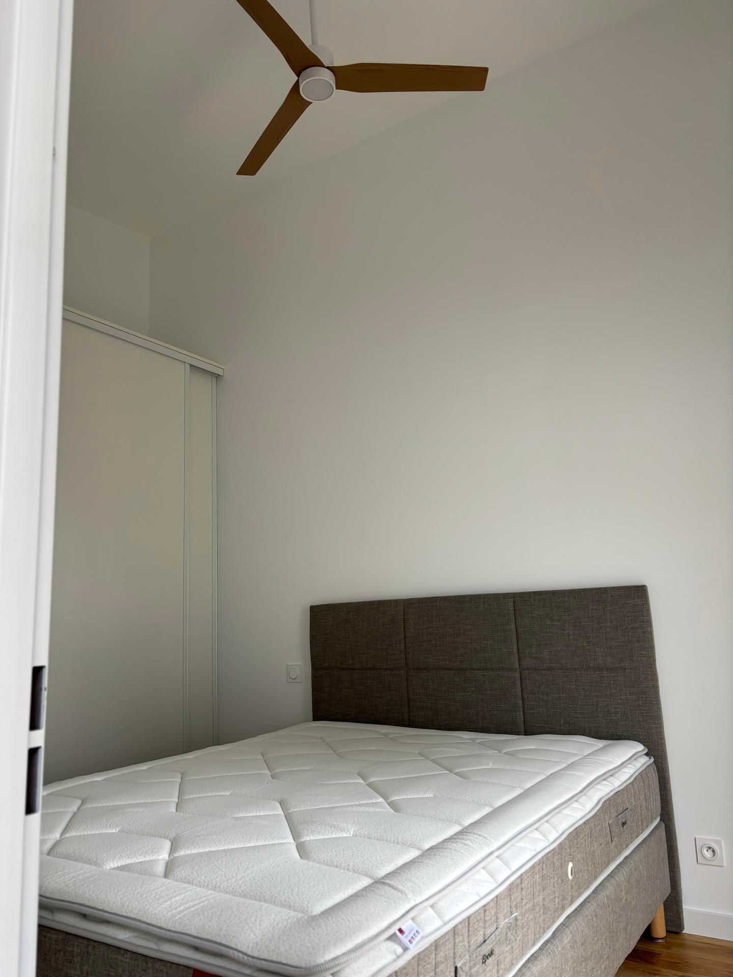 Petite chambre avec un lit, une tête de lit grise, du linge de lit blanc et un ventilateur de plafond.