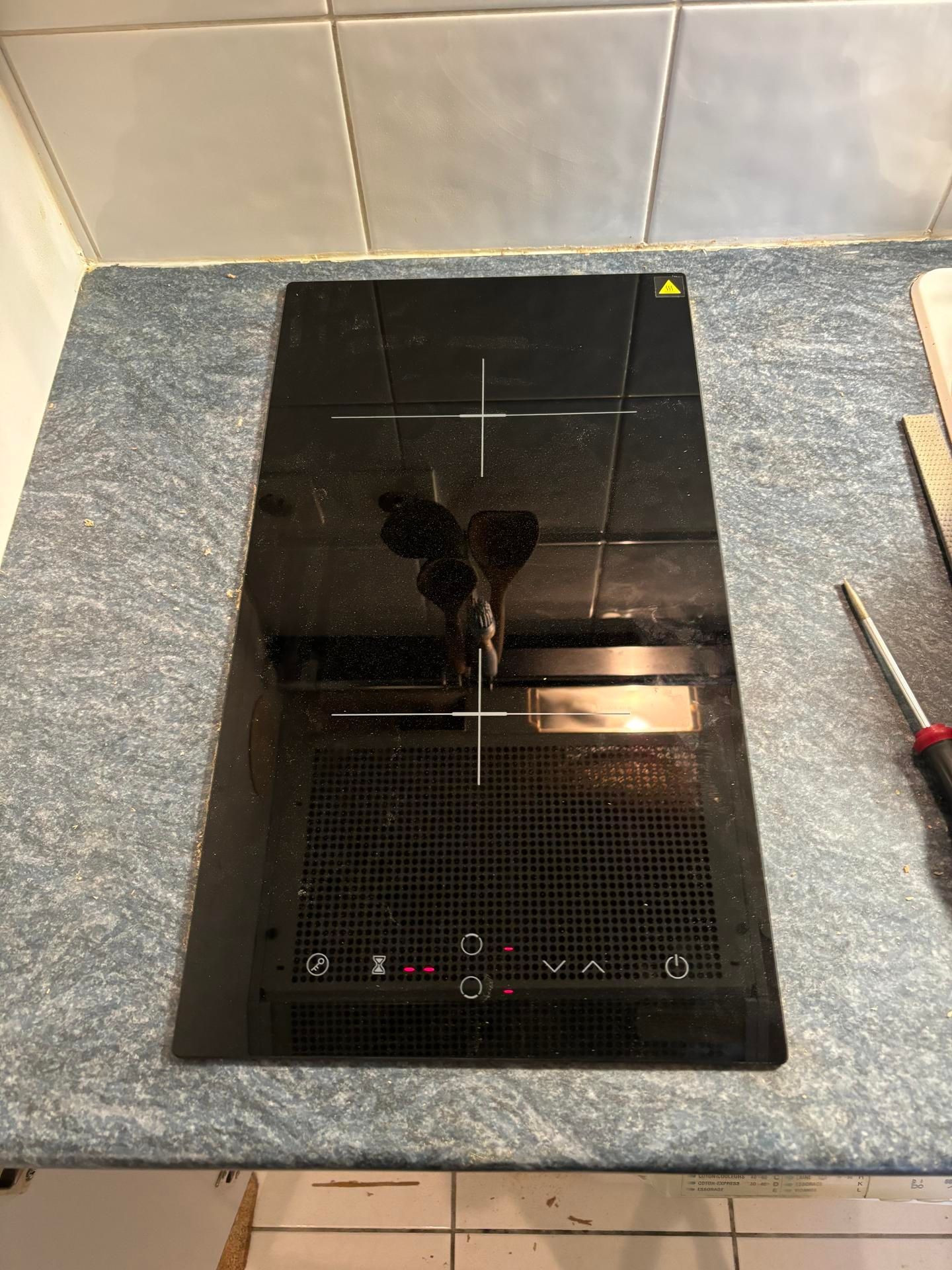 Plaque de cuisson à induction en verre noir sur un plan de travail gris avec crédence carrelée et un tournevis à proximité.