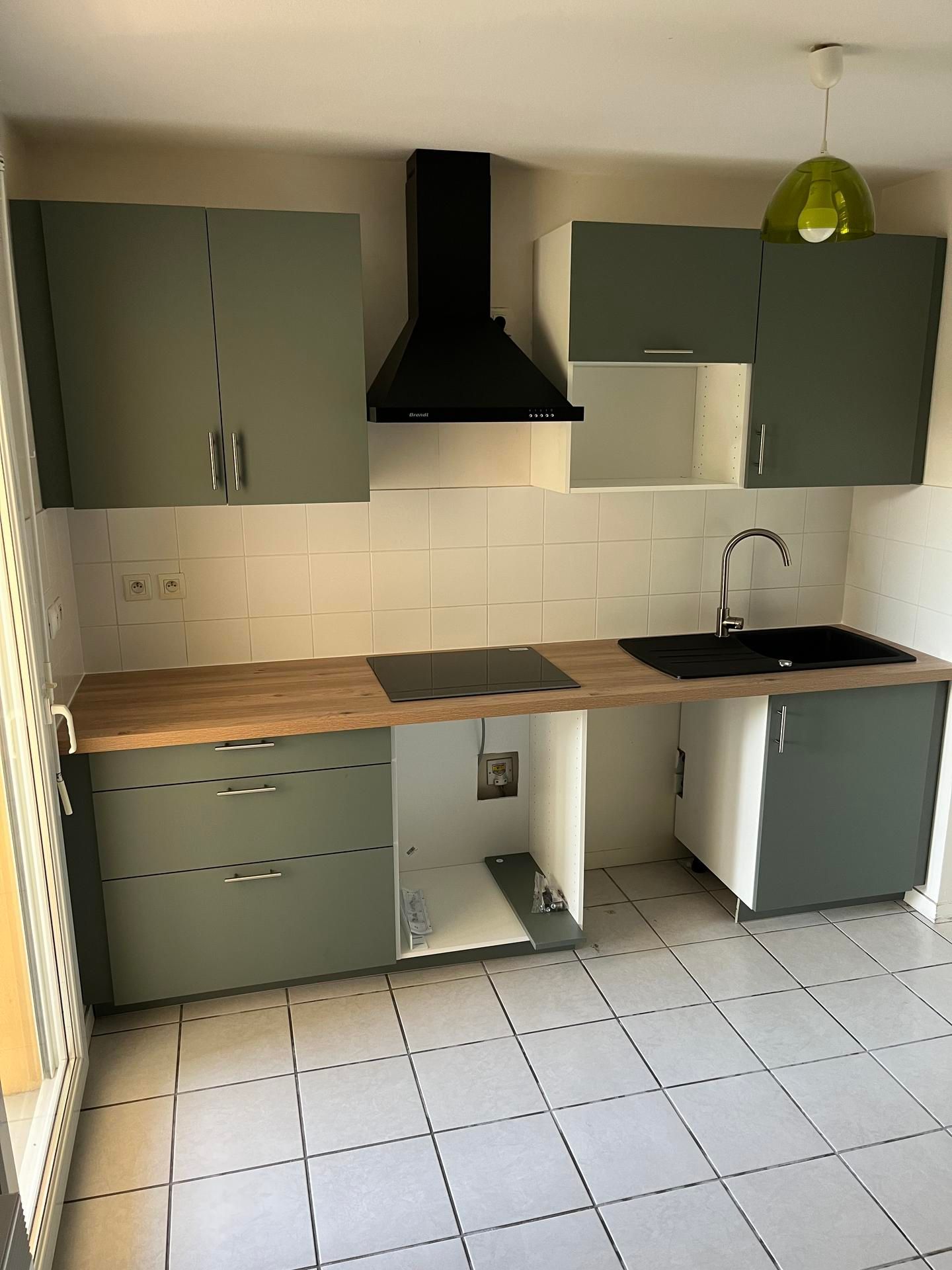 Petite cuisine avec placards verts, plan de travail en bois, évier noir, plaque de cuisson et sol carrelé.