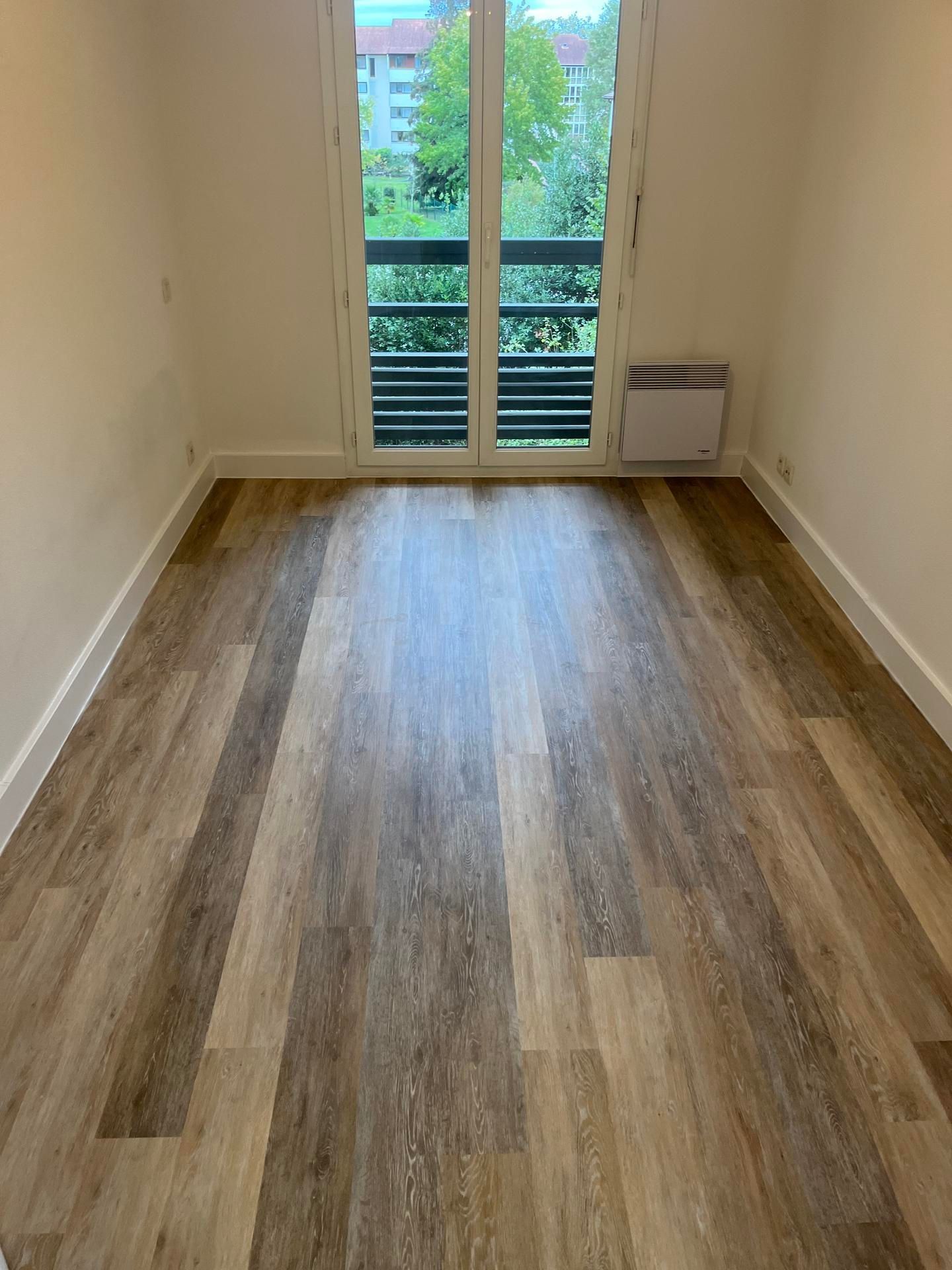 Pièce étroite et vide avec parquet, portes-fenêtres donnant sur un balcon et un petit radiateur mural.