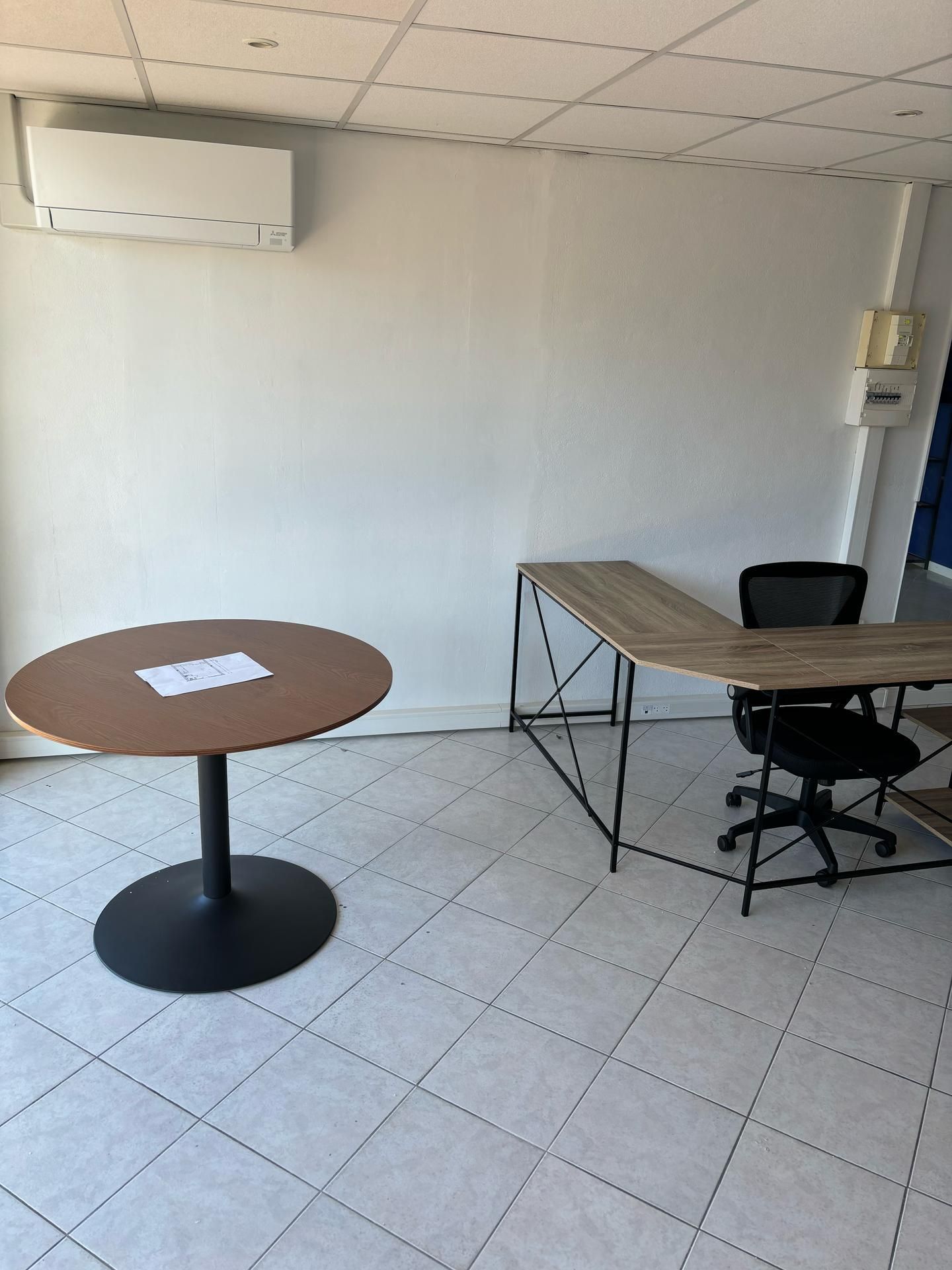 Petit bureau avec des bureaux, une chaise, un sol carrelé, une climatisation murale et une table ronde avec une feuille de papier.