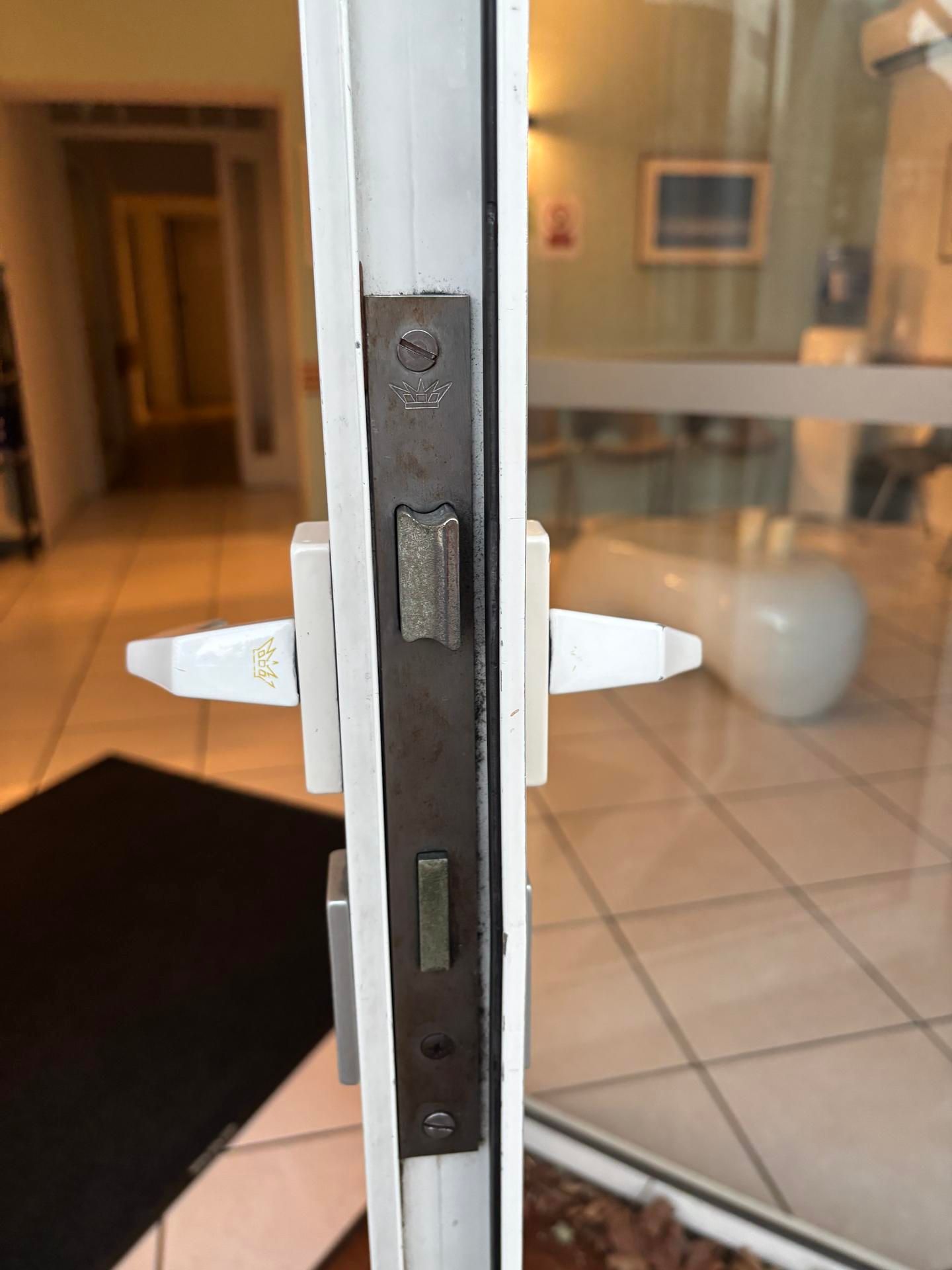 Gros plan sur le loquet et la gâche d'une porte vitrée, avec un couloir intérieur flou en arrière-plan.
