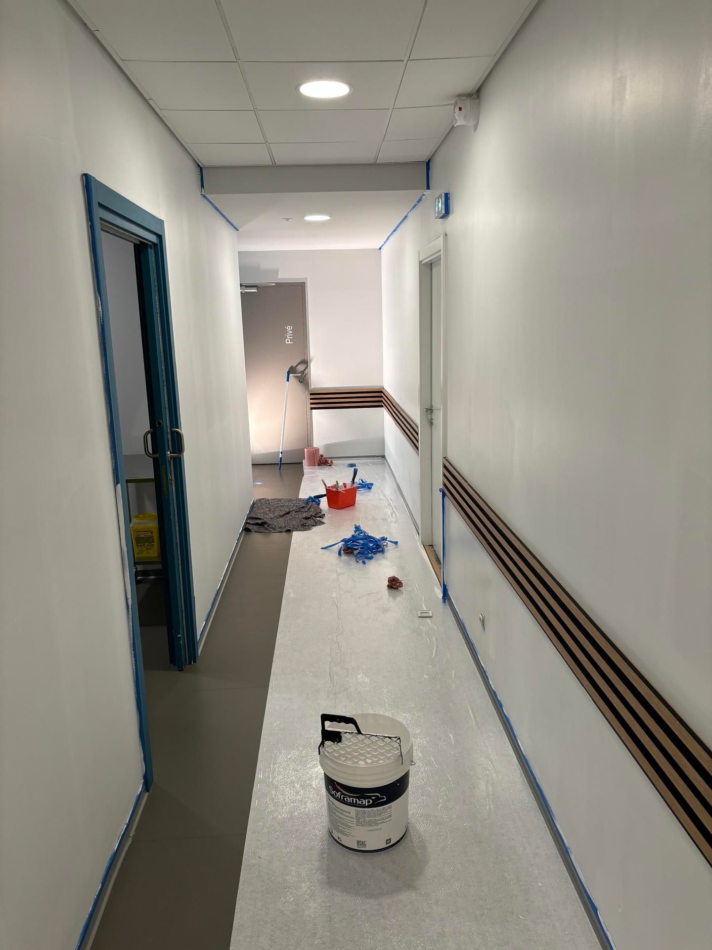 Couloir étroit en rénovation, avec des seaux de peinture et des débris au sol, les murs partiellement recouverts de plastique.