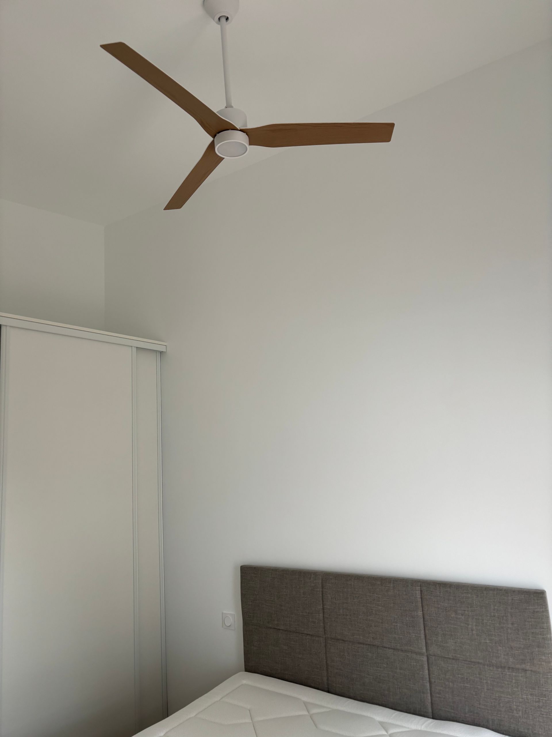Coin de chambre minimaliste avec un ventilateur de plafond marron, une armoire blanche et une tête de lit grise.