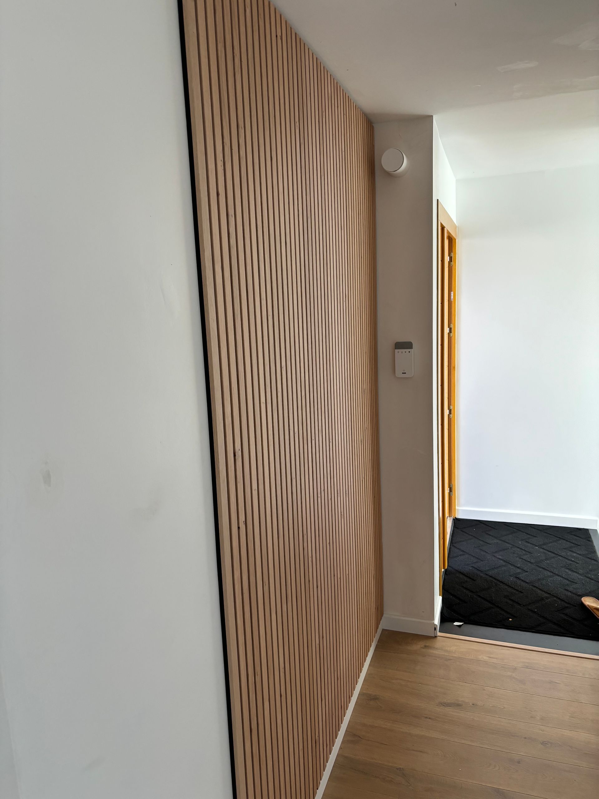 Couloir moderne avec mur lambrissé en lattes de bois, murs blancs, parquet clair et paillasson noir devant la porte.