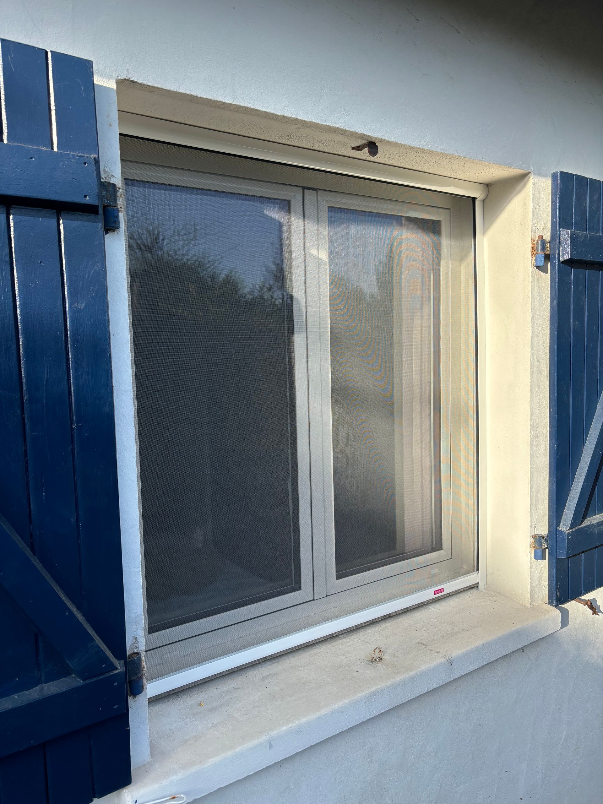 Fenêtre coulissante blanche fermée avec volets bleus sur un mur en stuc blanc.