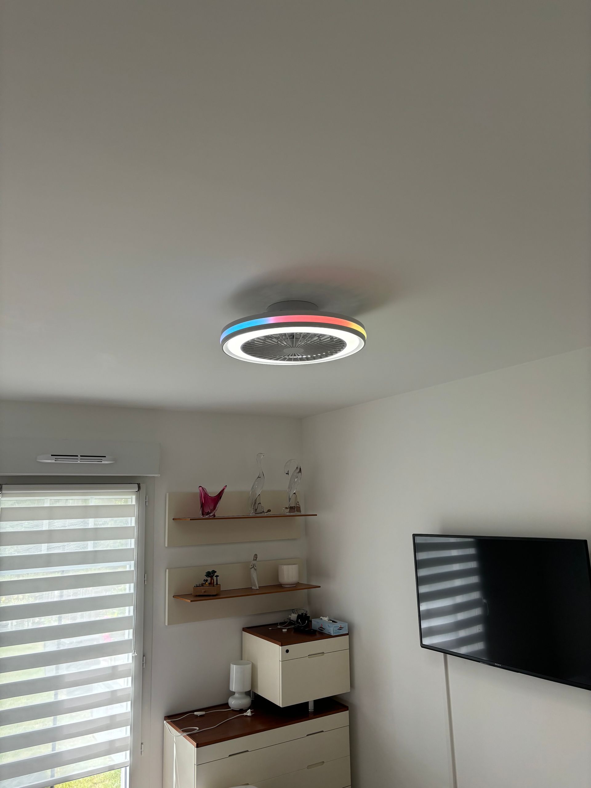 Petite pièce avec un plafonnier, une télévision fixée au mur, des étagères et une fenêtre avec des stores à rayures.