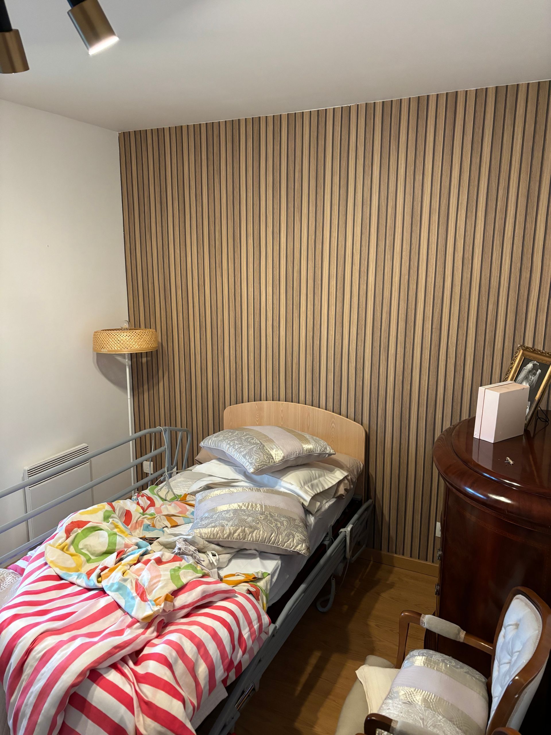 Petite chambre avec un lit simple, une couverture à rayures, du papier peint à rayures, un lampadaire et une table de chevet.