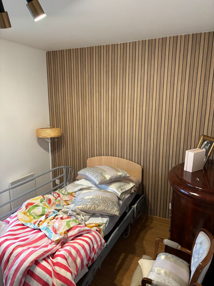 Petite chambre avec un lit simple, une couverture à rayures, du papier peint à rayures, un lampadaire et une table de chevet.