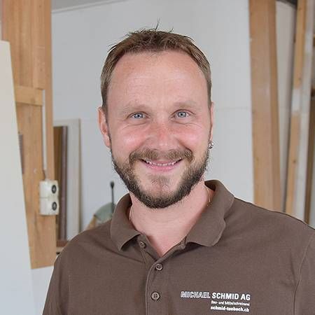 Ein Mann mit kurzem Bart lächelt in einer Werkstattumgebung in die Kamera.