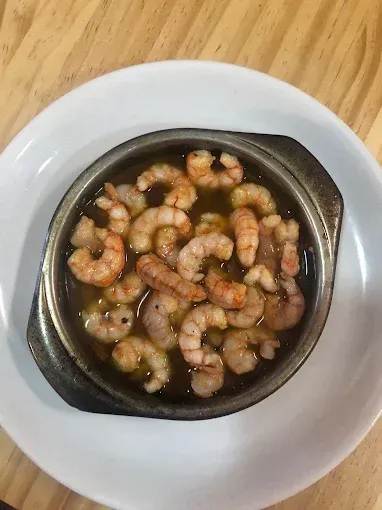 Camarones cocinados en aceite de oliva, servidos en un plato pequeño y oscuro sobre un plato blanco.